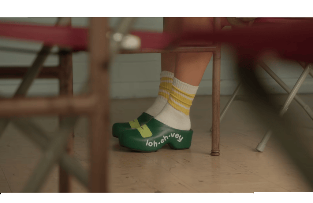 Close-up of a person's feet wearing green clogs with yellow accents and white socks with yellow stripes, seated between chairs in a classroom setting.