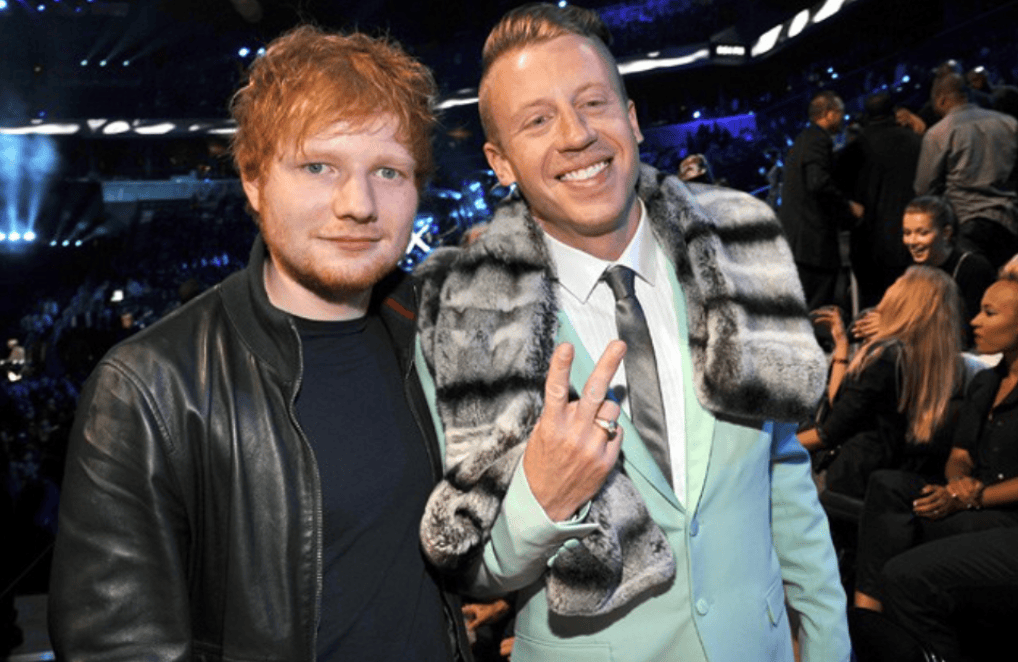 Two male musicians posing for a photo at an event, with one wearing a black leather jacket and the other in a light blue suit with a fur scarf.