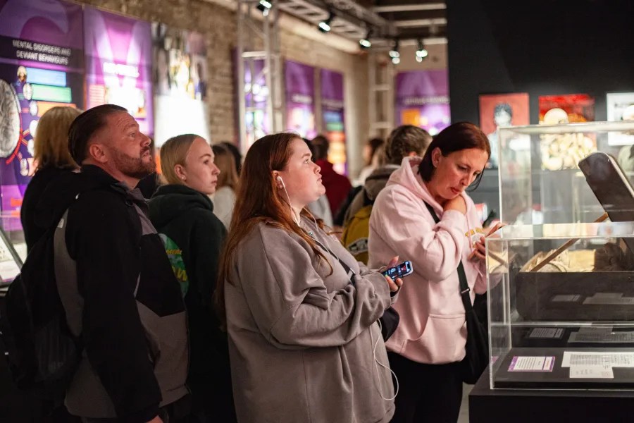 A group of people observing exhibits in a museum, with a focus on a display case. Some individuals are wearing headphones and using mobile devices while looking at the exhibits.
